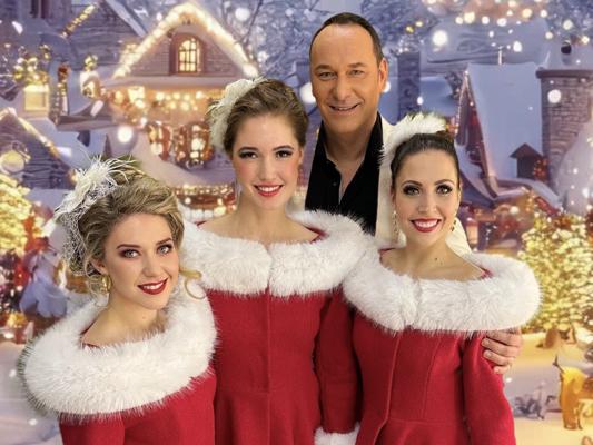 Drei Frauen in roten Kostümen mit weißem Fellkragen und ein Mann stehen vor einer festlich beleuchteten Winterkulisse.