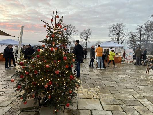 Ein geschmückter Weihnachtsbaum mit roten Kugeln und Lichterketten steht auf der Seeterrasse, Besucher an Marktständen.