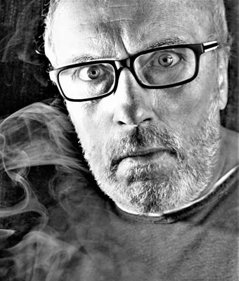 Ein Mann mit Brille und grauem Bart blickt direkt in die Kamera, während Rauch vor dunklem Hintergrund aufsteigt.