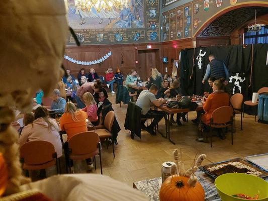 Im festlich geschmückten Saal sitzen Kinder und Erwachsene an Tischen, während ein Mann vor einer Halloween-Dekoration steht.