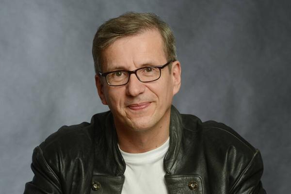 Ein Mann mit Brille, kurzem Haar und schwarzer Lederjacke blickt lächelnd vor einem grauen Studiohintergrund in die Kamera.