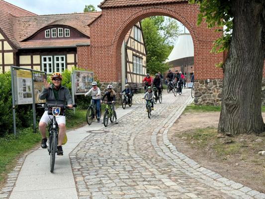 Mehrere Radfahrer fahren über einen Kopfsteinpflasterweg unter einem Backsteinbogen an historischen Gebäuden vorbei.