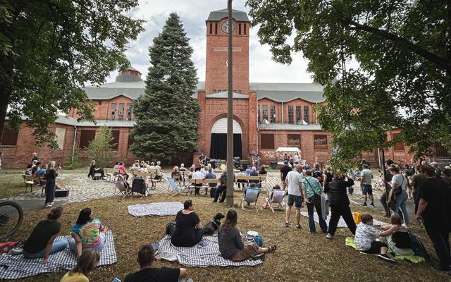 Vor dem historischen Backsteingebäude sitzen Besucher auf Decken und Stühlen, während mehrere Menschen Musik hören.