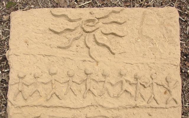 Ein Lehmrelief zeigt eine Sonne, geschwungene Linien und eine Reihe von stilisierten Figuren mit erhobenen Armen.
