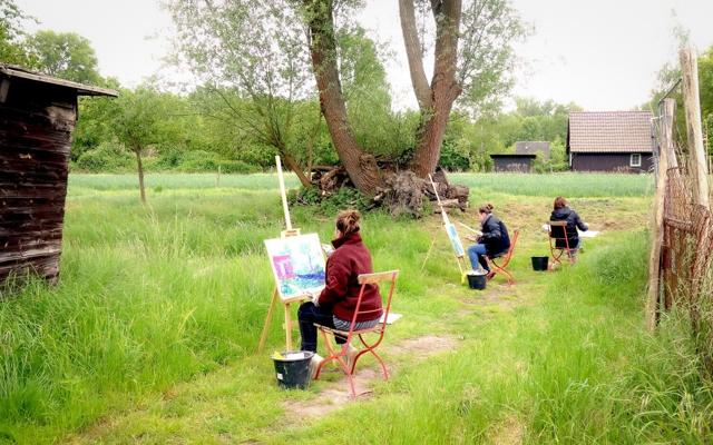 Drei Teilnehmende malen an Staffeleien auf einem Grasweg unter großen Bäumen, mit Farbkübeln und Häusern im Hintergrund.