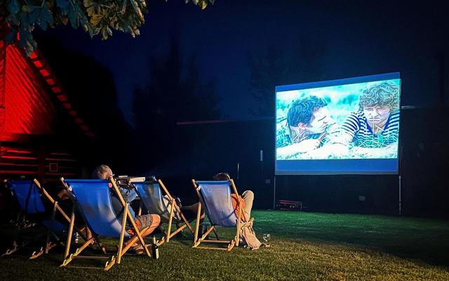 Bei Nacht sitzen mehrere Menschen in Liegestühlen auf einer Rasenfläche vor einer großen Leinwand mit einer Filmszene.