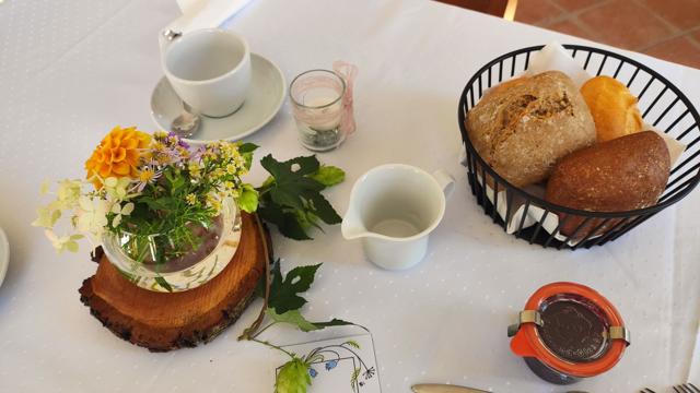 Auf dem weiß gedeckten Tisch stehen Tassen, ein Korb mit Brötchen, ein Blumenarrangement auf Holz und ein kleines Glas.