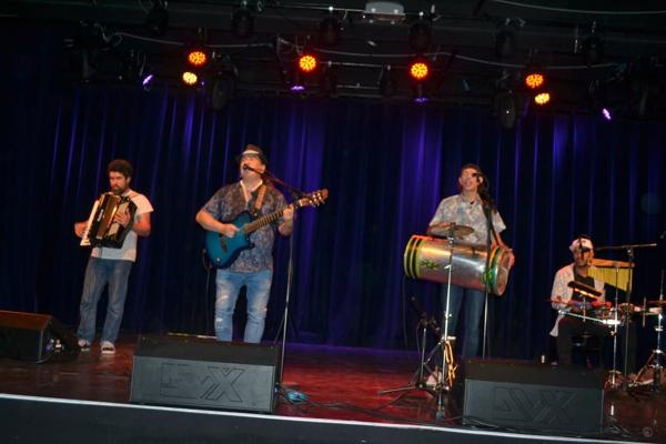 Vier Musiker spielen auf einer Bühne vor blauen Vorhängen, mit Akkordeon, Gitarre, Trommel und Schlagzeug.