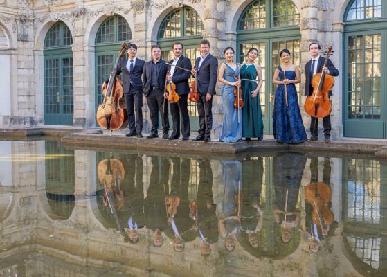 Mehrere Musiker stehen mit Streichinstrumenten vor einer historischen Fassade, deren Spiegelung sich im Wasser zeigt.