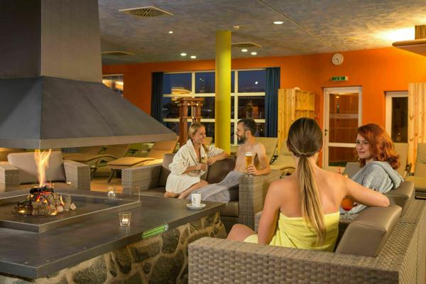 Mehrere Gäste sitzen in einer Saunalounge um einen offenen Kamin, trinken und unterhalten sich in warmem Licht.